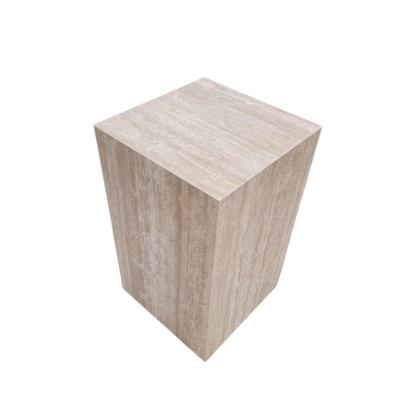 May include: A square, beige-colored pedestal with a textured, wood-grain appearance. The sides taper slightly, giving it a trapezoidal shape. The neutral color and simple design make it suitable for various decorative purposes.