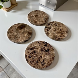 Può includere: Quattro sottobicchieri rotondi con un motivo marmorizzato marrone. I sottobicchieri sono disposti su una superficie bianca, mettendo in risalto il loro aspetto in pietra naturale. Questi sottobicchieri sono progettati per proteggere le superfici dalla condensa delle bevande.