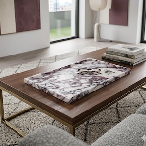 May include: A rectangular marble tray with a white and purple pattern sits on a dark brown wooden coffee table with a gold metal frame. The tray holds eyeglasses and keys. Books are stacked on the table.