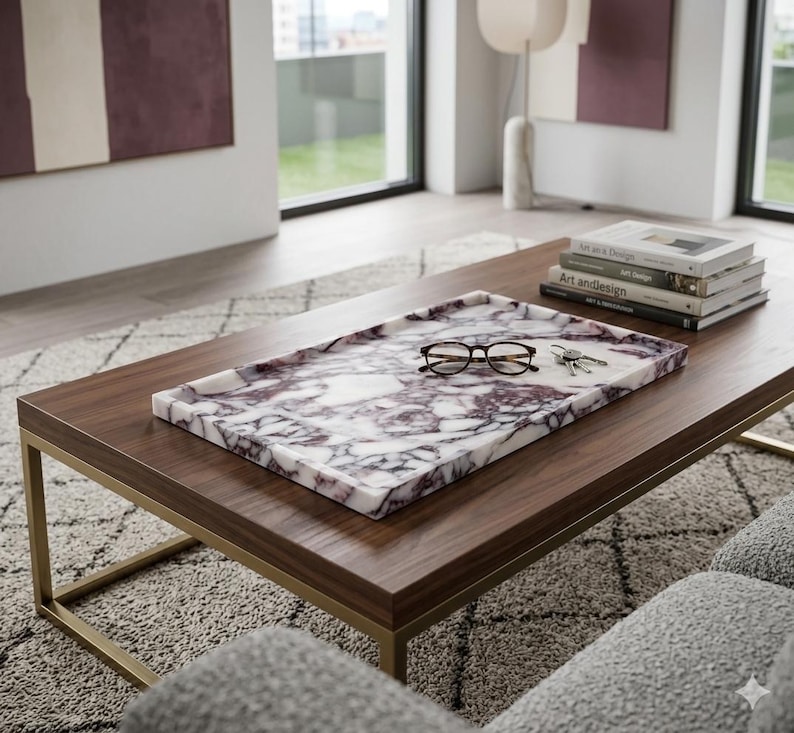 May include: A rectangular marble tray with a white and purple pattern sits on a dark brown wooden coffee table with a gold metal frame. The tray holds eyeglasses and keys. Books are stacked on the table.