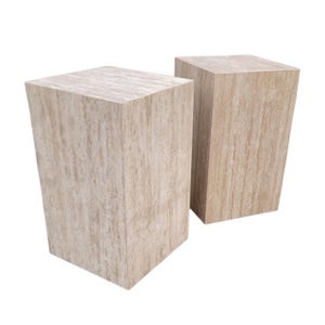 May include: Two light beige, rectangular pedestals with a natural, textured appearance. The pedestals have a trapezoidal shape, wider at the top and tapering towards the base. The neutral color and simple design suggest a modern aesthetic, suitable for displaying art or plants.