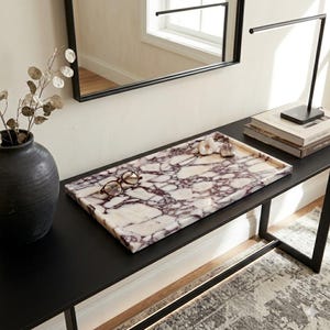 May include: A rectangular marble tray with a white and purple veined pattern. The tray holds a pair of eyeglasses and decorative flowers. The tray sits on a black table, with a black vase and a modern lamp in the background.