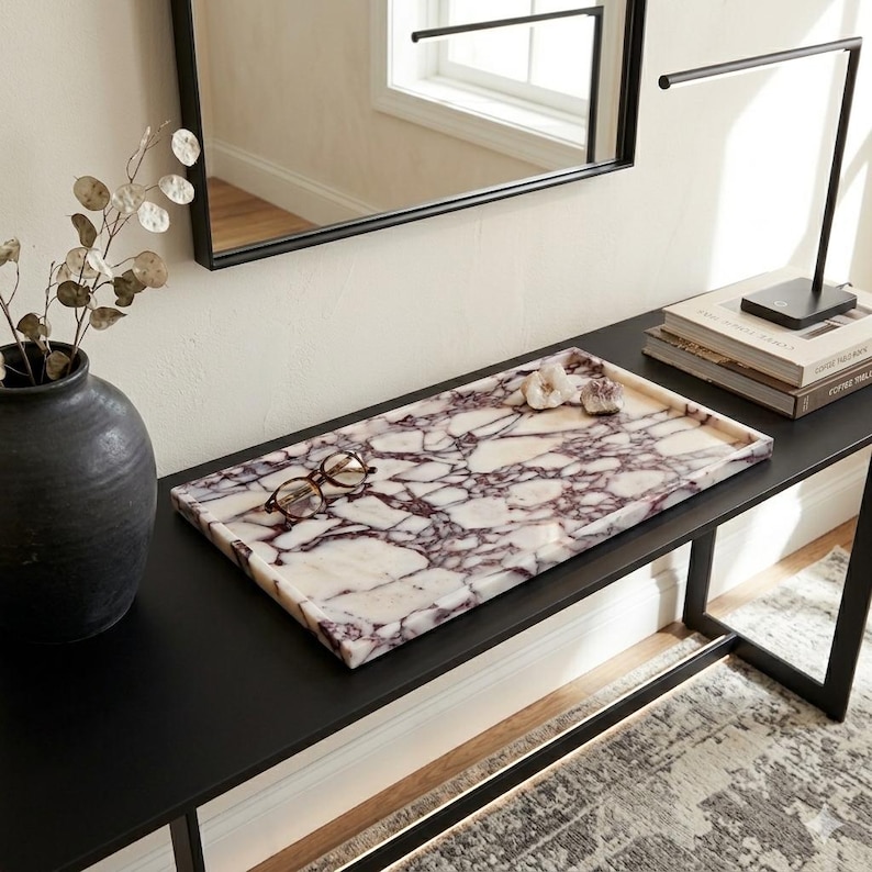 May include: A rectangular marble tray with a white and purple veined pattern. The tray holds a pair of eyeglasses and decorative flowers. The tray sits on a black table, with a black vase and a modern lamp in the background.