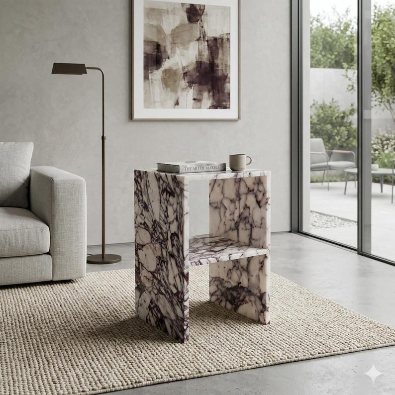 May include: A marble side table with a distinctive, open design. The table has a white base with dark purple veining. A book and a mug are on top. The table is in a modern living room setting.