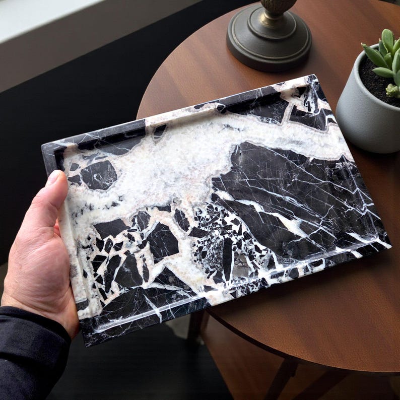 May include: A rectangular tray with a black and white marble pattern. The tray is held by a person's hand. The marble has a mix of black, white, and gray colors. The tray has a smooth, polished surface.