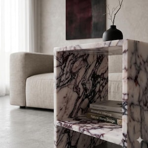 May include: A marble side table with a cube design, featuring a white and purple veined pattern. The table has two shelves and holds books. A black vase with a branch sits on top. The background includes a beige sofa and abstract art.