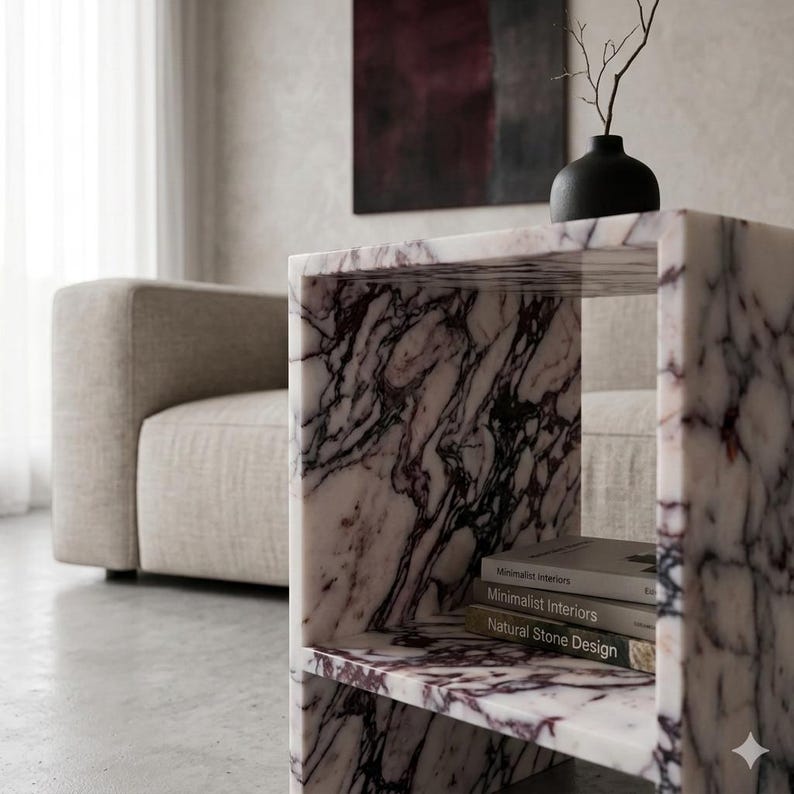 May include: A marble side table with a cube design, featuring a white and purple veined pattern. The table has two shelves and holds books. A black vase with a branch sits on top. The background includes a beige sofa and abstract art.