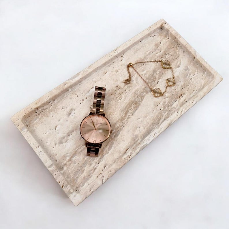 May include: A rectangular, beige travertine tray holds a rose gold watch and a delicate gold bracelet with clover-shaped charms. The watch has a round face with Roman numerals. The items are arranged on a white background.