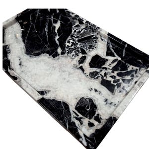 May include: A rectangular marble tray with a polished finish. The tray features a striking pattern of black and white veining, creating a natural, elegant aesthetic. The tray's edges are slightly raised, providing a defined border.