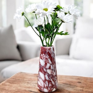 Pode incluir: Um vaso de mármore com um padrão vermelho e branco contém um buquê de margaridas brancas com centros amarelos e caules verdes. O vaso está sobre uma mesa de madeira, com um fundo branco desfocado.