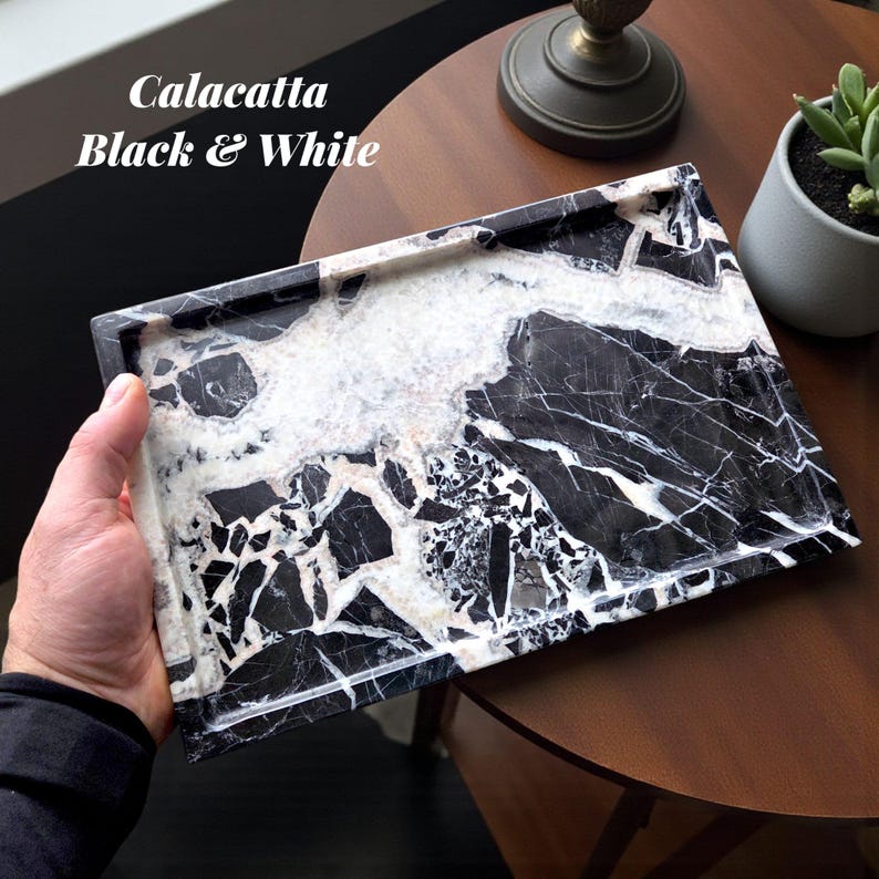 May include: A rectangular tray with a black and white Calacatta marble design. The tray has a polished finish and is held by a hand. The text "Calacatta Black & White" is visible in the upper left corner. The tray is on a wooden table.