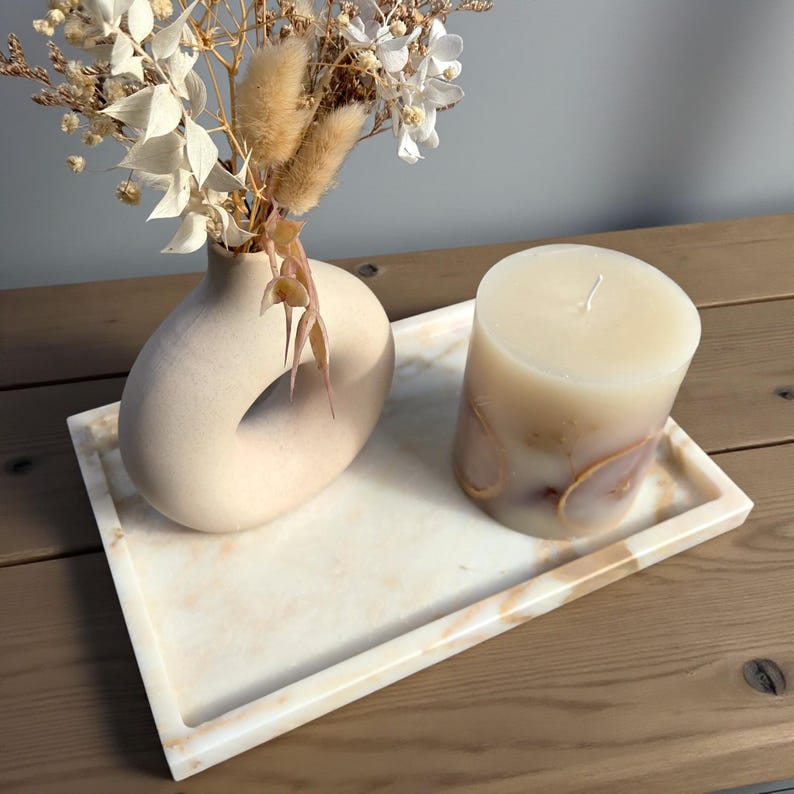 May include: A decorative tray holds a beige vase with dried flowers and a large pillar candle. The candle has a marbled design with brown and beige tones. The tray has a marble pattern with beige and light brown colors.