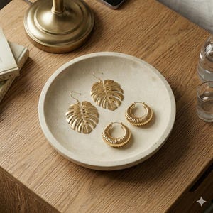 May include: A collection of four gold-toned earrings resting in a shallow, circular, off-white stone dish. The set includes two dangling earrings shaped like monstera leaves and two small, ribbed hoop earrings with a polished metallic finish.