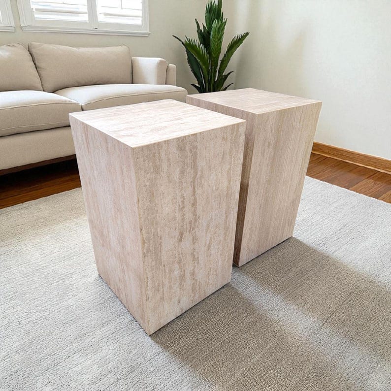 May include: Two light beige, rectangular side tables with a natural stone appearance. The tables are placed on a light gray rug, and have a slightly trapezoidal shape. The tables are next to each other.