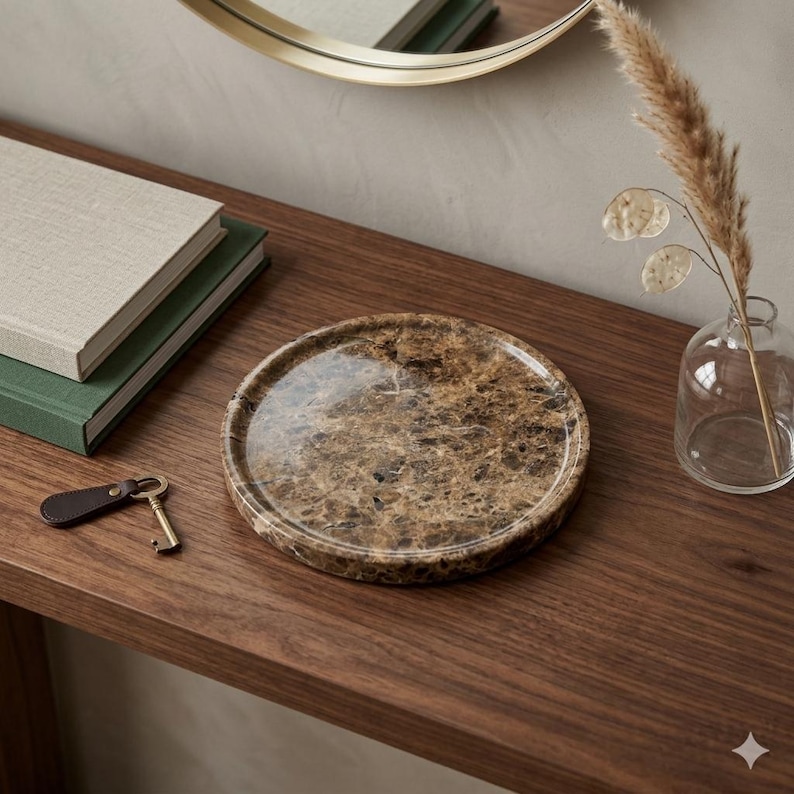 Puede incluir: Una bandeja redonda de m&aacute;rmol marr&oacute;n con una superficie pulida. La bandeja est&aacute; sobre una superficie de madera oscura, junto a un peque&ntilde;o jarr&oacute;n de cristal con flores secas, una pila de libros y una llave con un llavero de cuero.