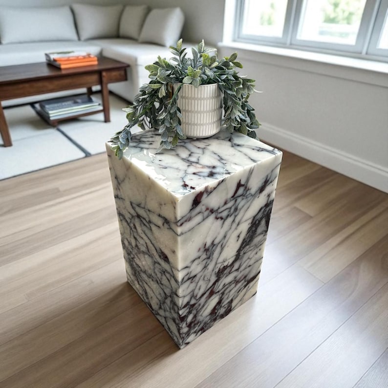 May include: A square marble side table with a white and dark purple veined pattern. A white ceramic pot with a green plant sits on top. The table is a decorative accent piece for a living room.