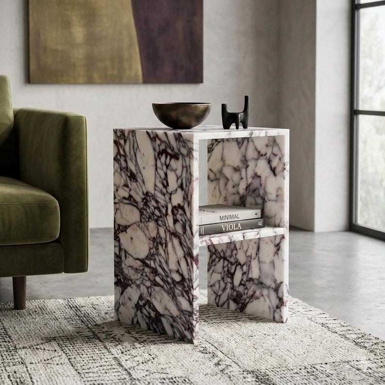 May include: A marble side table with a white and purple veined pattern. The table has a shelf with two books titled "Minimal" and "Viola". A dark metal bowl and a black cat sculpture sit on top. The table is 50 cm wide.