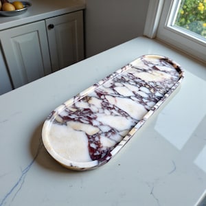 May include: An oblong marble tray with a polished finish. The tray has a white base with striking purple and grey veining. The edges are rounded, and the tray is set on a light-coloured countertop, suitable for serving or display.