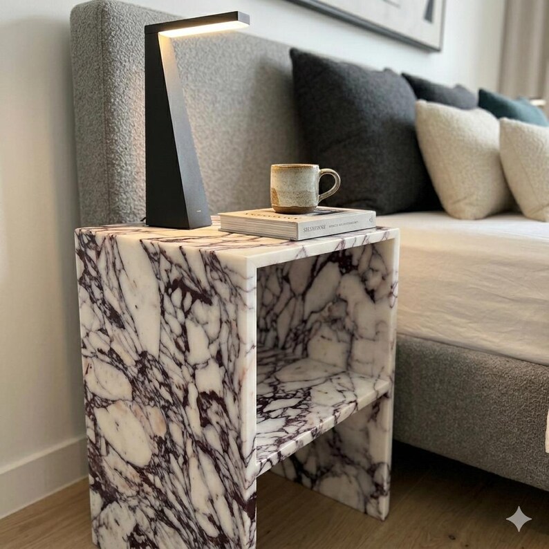 May include: A bedroom scene with a marble-effect side table, a black lamp, a mug, and a book. The table has a white and purple veined pattern. The bed has grey pillows and a grey headboard.