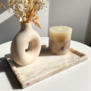 May include: A decorative arrangement featuring a beige vase with a circular opening, a pillar candle with dried fruit accents, and a marble tray. The vase holds dried flowers. The candle and vase sit on a rectangular marble tray with a light brown and white pattern.