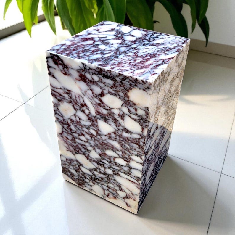 May include: A rectangular marble side table with a unique pattern of white, burgundy, and gray veining. The stone table is a cube shape and stands on a white tiled floor. The table is a decorative accent piece.