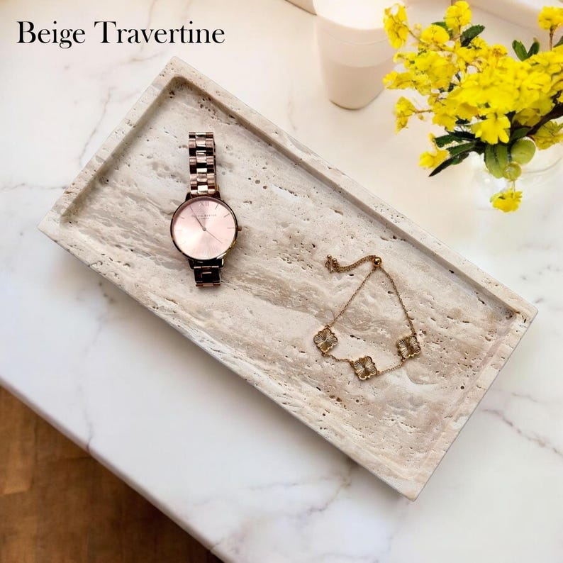 May include: Beige travertine rectangular tray with a rose gold watch and a gold bracelet with clover-shaped charms. The tray is on a white marble surface. The text "Beige Travertine" is at the top.