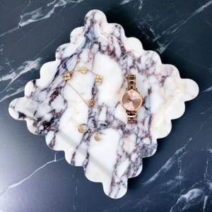 May include: A marble tray with a scalloped edge, displaying a gold watch, necklace, and earrings. The tray has a white and purple veined pattern. The watch features a rose gold band and face. The jewellery includes a clover necklace and small hoop earrings.