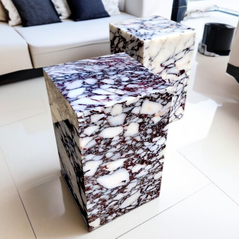May include: Two square marble side tables. The tables are a mix of white, burgundy, and dark purple. The tables are made of marble and have a polished finish. The tables are cube-shaped and are placed on a white tiled floor.