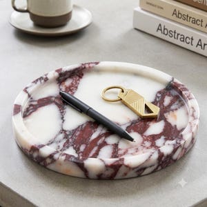 May include: A round, white marble tray with deep red veining. Resting on the tray is a matte black ballpoint pen and a gold-toned metal keychain featuring a geometric chevron pattern and a triangular cutout at the bottom edge of the tag.