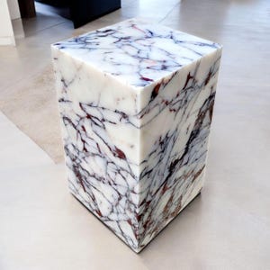 Pode incluir: Uma mesa lateral cúbica de mármore branco com veios vermelhos escuros e cinzentos. A mesa de pedra tem um acabamento polido e um design geométrico. O cubo é uma peça de destaque moderna para uma sala de estar ou quarto.