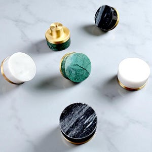 May include: A selection of cabinet knobs in various designs. The knobs feature white, green, and black marble, complemented by gold-coloured metal. The knobs are round and cylindrical, offering a contemporary look for furniture.