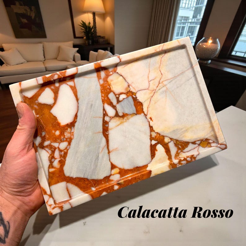 May include: A rectangular Calacatta Rosso marble tray with a polished finish. The tray features a pattern of white, orange, and gray veining. The text "Calacatta Rosso" is printed in black at the bottom.