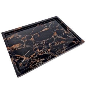 Puede incluir: Una bandeja rectangular de m&aacute;rmol negro con borde elevado. La bandeja presenta un llamativo patr&oacute;n de vetas marrones y blancas en toda la superficie pulida. Esta bandeja decorativa es ideal para servir o exhibir art&iacute;culos.