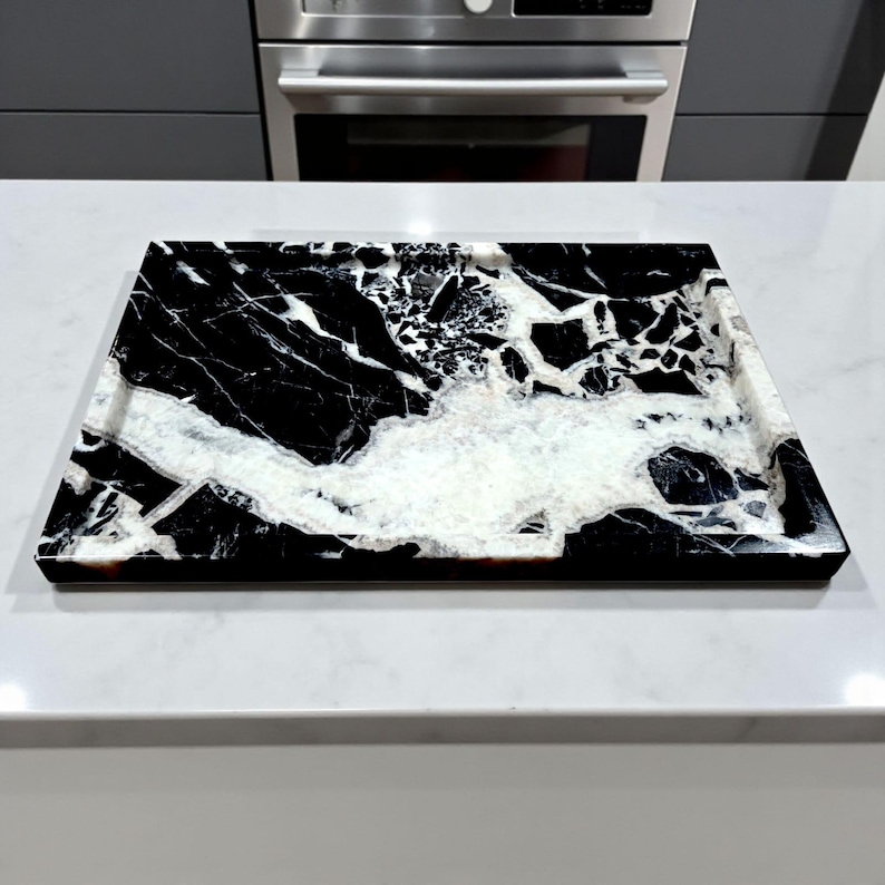May include: Rectangular black and white marble tray. The tray features a striking pattern of black veining against a white background. The tray is set on a white countertop, with a stainless steel appliance in the background.