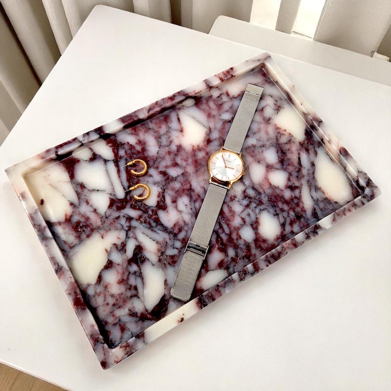 May include: A rectangular tray with a marbled design in shades of burgundy and white. A gold-toned watch with a silver band and two gold hoop earrings rest on the tray. The tray is a decorative item for jewelry and accessories.
