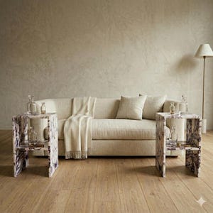 May include: A neutral-toned sofa with a cream throw and cushions. Two side tables with a marble effect and glass bottles flank the sofa. The tables have a purple and white marble pattern. A floor lamp is also visible.