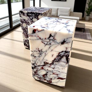 May include: Two marble cube side tables. The tables have a white base color with dark purple and red veining throughout. The tables are rectangular and have a modern design. The tables are placed on a light wood floor.