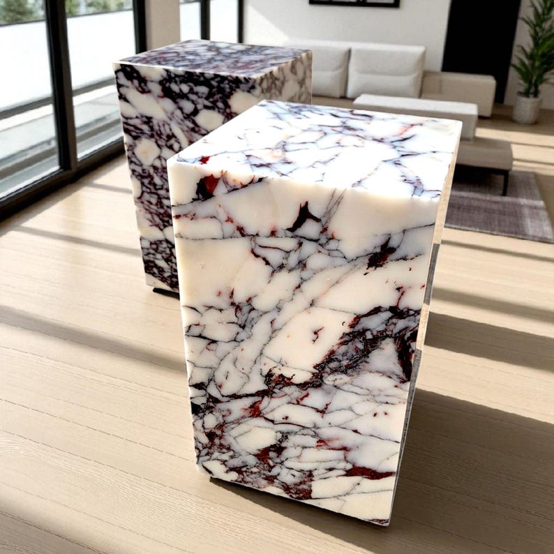 May include: Two marble cube side tables. The tables have a white base color with dark purple and red veining throughout. The tables are rectangular and have a modern design. The tables are placed on a light wood floor.
