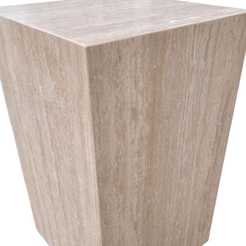 May include: A light beige, trapezoid-shaped pedestal or side table. The stone-like material has a natural, veined pattern. The top surface is flat, and the sides slope inward, creating a modern, geometric design. This accent table is suitable for home decor.