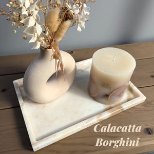 May include: A decorative tray with a beige marble pattern holds a beige vase with dried flowers and a large pillar candle. The candle has a marbled design and sits on a small marble tray. The text "Calacatta Borghini" is visible.