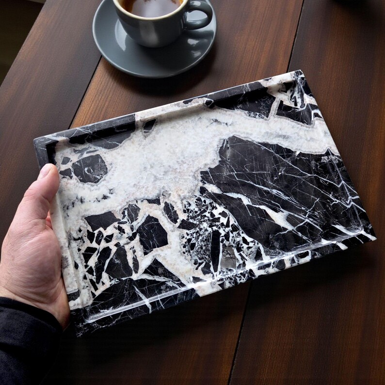 May include: Rectangular black and white marble tray with a unique veining pattern. The tray has a polished finish and is held by a person's hand. A gray teacup and saucer are in the background on a dark wooden surface.