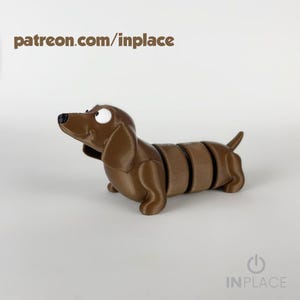 Może przedstawiać: Brązowa figurka psa jamnika z segmentowanym korpusem, czarnym nosem i dużym białym okiem. Tekst "patreon.com/inplace" znajduje się u góry. Figurka stoi na białym tle. Logo IN PLACE znajduje się na dole.