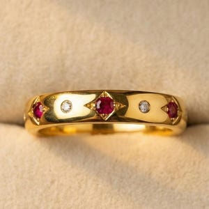 May include: A gold band ring featuring a repeating pattern of alternating round red rubies and clear white diamonds set within small, star-shaped metal indentations along the center of the polished yellow gold surface.