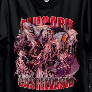 Puede incluir: Camiseta negra con un gráfico de personajes de Castlevania, con el texto "ALUCARD" y "CASTLEVANIA" en rojo. La camiseta está colgada en una percha negra contra una pared de ladrillos oscuros.