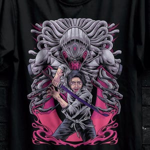 May include: Black t-shirt featuring a graphic of a warrior with a sword facing a monster. The design includes shades of pink, gray, and white. The shirt is hanging on a black hanger against a dark brick wall.