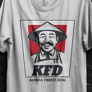 May include: A light gray t-shirt featuring a parody design. The design includes a smiling man in a conical hat, a red background, and the text "KFD KOREA FRIED DOG". The shirt is hanging on a black hanger.