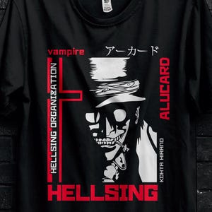 Può includere: T-shirt nera con grafica di un vampiro e le scritte rosse "HELLSING" e "ALUCARD". Il design include anche testo giapponese e il logo "HELLSING ORGANIZATION". La maglietta è appesa a una gruccia nera.