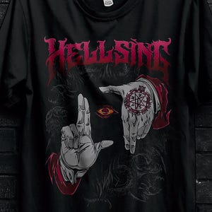 Può includere: T-shirt nero con la scritta "HELLSING" in lettere gotiche rosse. La maglietta presenta una grafica di mani che incorniciano un occhio e un simbolo. Il logo Stitch Aura si trova nell'angolo in alto a sinistra.