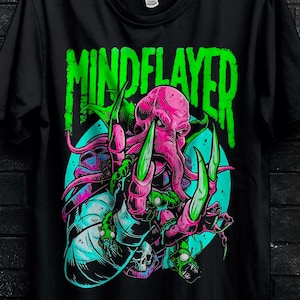 Puede incluir: Camiseta negra con un gráfico colorido de una criatura rosa con forma de pulpo con garras verdes y la palabra "MINDELAYER" en verde. El diseño incluye un fondo turquesa y detalles de calaveras. La camiseta está colgada en una percha negra.