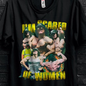 Peut inclure: T-shirt noir avec un motif d'hommes musclés et le texte jaune "I'M SCARED OF WOMEN". Le t-shirt est suspendu à un cintre noir devant un mur de briques sombres.